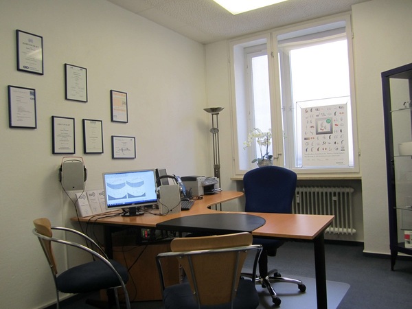 Modernes Hörakustiker-Beratungszimmer mit Schreibtisch, Computer und Zertifikaten an der Wand.